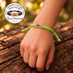 May include: A bright lime green woven cord bracelet featuring a rectangular wooden centerpiece engraved with a fish graphic. The bracelet has adjustable knotted ends and a circular logo overlay in the corner with the text Rowan Threadworks.
