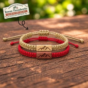 Könnte beinhalten: Zwei verstellbare Kordelarmbänder mit rechteckigen Holzperlen, die mit einem Gebirgsmotiv graviert sind. Ein Armband besteht aus goldfarbener Kordel, das andere aus roter Kordel. Ein Etikett trägt die Aufschrift Rowan Threadworks.