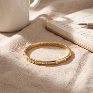 May include: A gold-toned metal bangle bracelet featuring a series of embedded clear stones in various shapes, including round, marquise, and diamond cuts, arranged in a repeating pattern along the outer surface of the circular band.