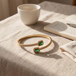 Könnte beinhalten: Ein goldfarbenes, offenes Armband mit zwei rechteckigen, smaragdgrünen Steinen an den Enden, die von kleinen, klaren Pavé-Kristallen entlang des Bandes und der Steinfassungen umgeben sind, was für ein helles, metallisches Aussehen sorgt.