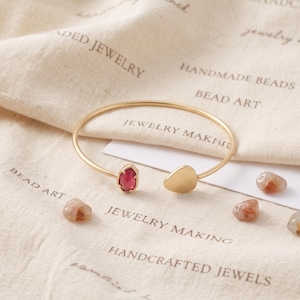 May include: A gold-toned open cuff bracelet featuring a single oval-shaped red gemstone on one end and a smooth, flat gold-toned teardrop-shaped charm on the other end. The bracelet is thin and circular, designed to be worn around the wrist.