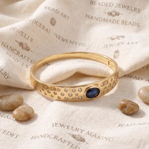 May include: A gold-toned hinged bangle bracelet featuring a polished finish, a central oval-shaped deep blue gemstone, and a scattered pattern of small, clear round crystals embedded across the front surface of the metal band.