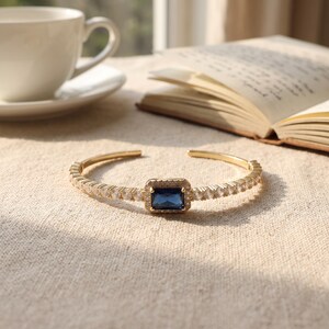 Op de afbeelding: Een goudkleurige armband met een centrale rechthoekige diepblauwe edelsteen, omringd door een halo van kleine heldere steentjes. De band is aan beide zijden bezet met een doorlopende rij heldere steentjes voor een symmetrisch ontwerp.