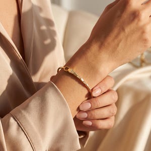 May include: A gold bangle bracelet featuring a slender band and a decorative focal point composed of three small, triangular segments inlaid with tiny clear stones. The bracelet has a polished metallic finish and a simple, open-ended design.