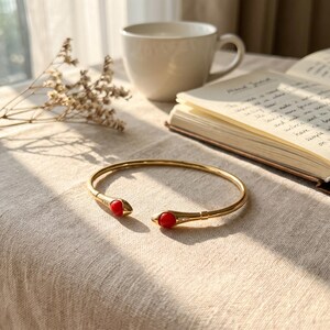 May include: A gold-toned open cuff bracelet featuring two rounded, bright red stone accents at each terminal end. The metal band is smooth and polished, with a small, subtle decorative detail near each of the red stone settings.