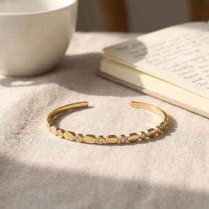 May include: A gold-toned open cuff bracelet featuring a repeating pattern of smooth oval metal links alternating with small, round, clear, faceted stones set into the band, creating a delicate and symmetrical design across the entire piece.