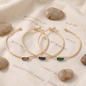 May include: Three gold-tone metal cuff bracelets, each featuring a single rectangular baguette-cut gemstone. The stones are purple, dark blue, and green. Each bracelet has a thin, open-ended band with a small spherical bead at each terminal end.