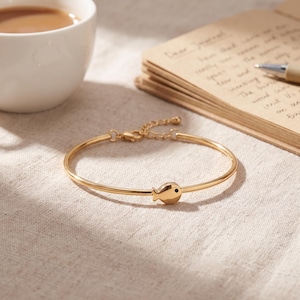 May include: A gold-toned metal bangle bracelet featuring a small, polished fish charm at the center. The fish charm has a single tiny black stone for an eye. The bracelet includes a thin chain extension with a lobster clasp closure for adjustment.