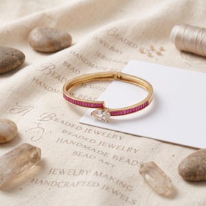 Op de afbeelding: Een goudkleurige scharnierende armband met een rij rechthoekige magentakleurige stenen langs de band en een enkele peervormige doorzichtige steen bij de opening in het midden. De armband wordt getoond op een witte kaart.