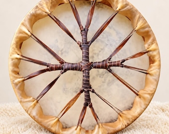 May include: A round frame drum featuring a translucent, light-colored animal hide head stretched over a circular wooden frame. Dark brown leather lacing crisscrosses the back, forming a central cross-like structure with wrapped details.