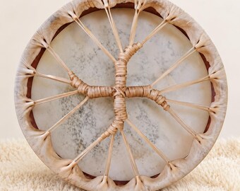 May include: A circular frame drum featuring a light-colored, mottled animal hide drumhead stretched over a wooden hoop. The drumhead is secured by thin, light-tan leather lacing that weaves into a central cross-shaped design.