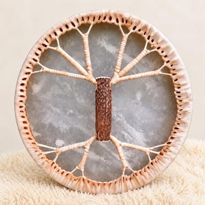 May include: A circular frame drum featuring a translucent gray drumhead. The frame is wrapped in light beige leather lacing that extends across the drumhead to form a central, textured brown wooden handle resembling a tree trunk.