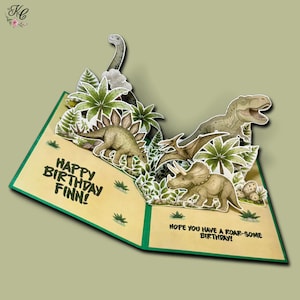 May include: A pop-up birthday card featuring a prehistoric dinosaur scene with a Stegosaurus, a T-Rex, a Pterodactyl, and a Triceratops among green foliage. The text reads: Happy Birthday Finn! Hope you have a roar-some birthday! The card is cream-coloured.