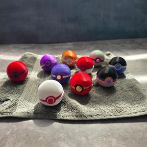 May include: A collection of eleven spherical toys resembling Pokeballs in various colours including red, white, purple, orange, and black. Each sphere features a central circular button detail and a horizontal band encircling the middle of the object.
