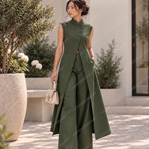 Op de afbeelding: Een olijfgroene mouwloze tuniek met een mandarijnkraag en drie knopen aan de zijkant, gecombineerd met een bijpassende broek met wijde pijpen. De tuniek heeft een lang, aan de voorzijde open ontwerp dat tot op de knieën valt. Een kleine beige handtas.