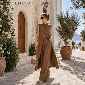Può includere: Un completo marrone composto da un gilet lungo senza maniche e pantaloni ampi abbinati. Il gilet presenta un colletto alto e uno spacco frontale. L'outfit è abbinato a una piccola borsa a tracolla beige con catena dorata. Testo ETHNIA STORE ART.