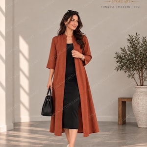 May include: A long, rust-coloured open-front duster coat with three-quarter sleeves and a mandarin collar, worn over a simple black midi dress. The outfit is paired with white trainers and a black structured handbag, creating a layered look.