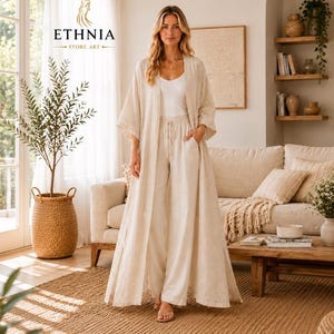 May include: A beige long-sleeved open-front duster cardigan worn over a white tank top and matching beige wide-leg trousers with a drawstring waist. The outfit features a light, textured fabric and a loose, flowing silhouette.
