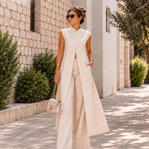 Könnte beinhalten: Eine ärmellose, lange beigefarbene Tunika mit Stehkragen und einem Schlitz vorne, kombiniert mit einer passenden weiten Hose. Das Outfit wird durch eine kleine beigefarbene Handtasche mit goldener Kette, goldene Armreifen und eine dunkle Sonnenbrille ergänzt.