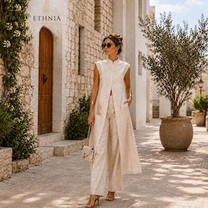 May include: A beige sleeveless long vest with a front button closure paired with matching wide-leg trousers. The outfit is worn with a small beige shoulder bag featuring a gold chain strap and gold-toned hardware, plus brown heeled sandals.