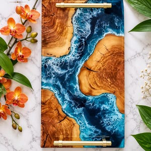 May include: A rectangular serving tray made of natural wood with a live edge and a central river-like inlay of blue and white resin. The tray features two polished gold-tone metal handles attached to the top and bottom edges of the piece.