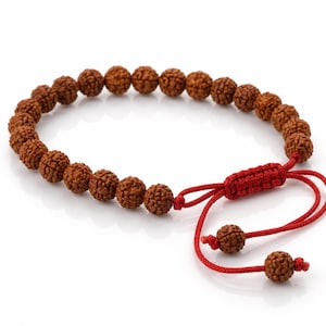 May include: A bracelet featuring a series of round, textured brown rudraksha beads strung on a vibrant red cord. The design includes an adjustable sliding knot closure with two dangling red cord ends, each finished with a single matching brown bead.