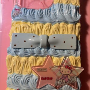 Peut inclure: Une coque de téléphone transparente décorée de textures de crème fouettée jaune et bleu clair. Elle présente un nœud bleu clair à pois marron, deux breloques boutons à carreaux bleus et une étoile avec le motif et le texte Hello Kitty.