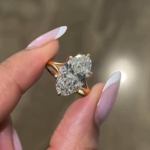 May include: A gold ring featuring two large, clear, marquise-cut stones set side-by-side in a prong setting. The band is thin and polished gold, providing a bright contrast to the faceted, brilliant-cut stones that catch the light.