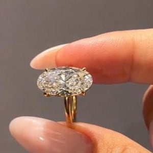 May include: A gold ring featuring a large, oval-cut, clear faceted gemstone set in a four-prong setting. The band is thin and polished gold, providing a simple and clean foundation for the prominent, horizontally oriented oval stone.