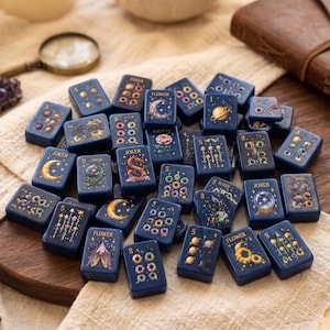 Può includere: Una collezione di tessere rettangolari blu scuro con illustrazioni celesti e floreali in oro e colori vivaci. Ogni tessera presenta motivi unici tra cui lune, pianeti, fiori e il testo Joker, Flower o vari numeri.