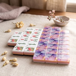 Op de afbeelding: Een set rechthoekige mahjongtegels gerangschikt in een raster. Het ene deel bevat witte tegels met groene bamboe, rode karakters en kleurrijke vogelillustraties. Het andere deel bestaat uit tegels met een paars, roze en wit marmerpatroon.