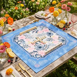 A floral-patterned game board with the word "JOKER" displayed on it, surrounded by various tableware and flowers on a wooden table.