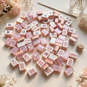 Op de afbeelding: Een verzameling rechthoekige mahjongtegels met een mix van witte oppervlakken met rode of zwarte symbolen en doorschijnende, met roze glitter gevulde achterkanten. De tegels liggen verspreid over een oppervlak met een houten dobbelsteen.