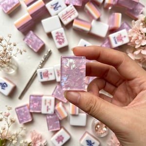 Op de afbeelding: Een rechthoekige mahjongtegel met een doorschijnende, iriserende roze afwerking met ingebedde glinsterende vlokken. De tegel wordt vastgehouden voor een verzameling soortgelijke mahjongtegels met witte basissen en roze, oranje of blauwe symbolen.