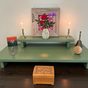 May include: A green wooden altar with two levels, featuring a small wooden box, a white vase with pink and purple flowers, two brass candle holders with lit candles, a small painting, a brass bell, and a decorative stone. The altar is decorated with a gold sun symbol.