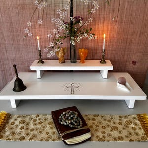 May include: A white altar table with a cross design, holding a bell, a book with rosary beads, and a decorative runner with gold fringe. A smaller shelf above holds candles, vases, and flowers. The background is a pink floral design.