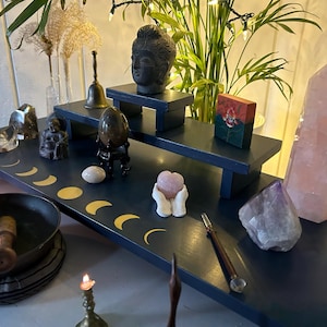 May include: A dark blue tiered display shelf with gold crescent moon accents, holding various crystals, a Buddha head, and a singing bowl. A large pink quartz crystal and amethyst are also displayed.