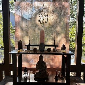 May include: A dark wooden altar table with a seated Buddha statue, crystals, and candles. A large pink crystal stands behind the table, with a decorative hanging star above. The background features a sheer curtain and sunlight.