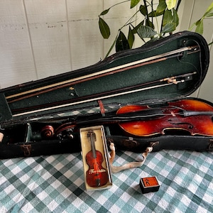 May include: A vintage violin with a dark brown finish, resting in an open black case with green lining. Two bows are inside the case. A miniature violin in a box and a small black box are also present.