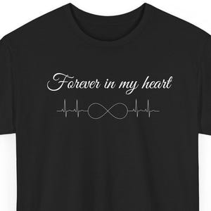 Può includere: Una t-shirt nera a maniche corte con il testo bianco "Forever in my heart" in carattere corsivo, posizionato sopra un disegno grafico a linee bianche di un battito cardiaco combinato con un simbolo di infinito centrato sul petto.