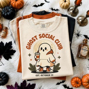 Può includere: Una t-shirt color crema con la grafica di un fantasma che indossa una felpa con cappuccio e scarpe da ginnastica mentre tiene in mano una tazza di caffè. Il testo recita GHOST SOCIAL CLUB e EST. OCTOBER 31. Il design include luna e stelle.