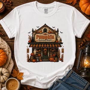 Può includere: Una t-shirt bianca a maniche corte con un'illustrazione grafica di una vetrina con la scritta Pumpkin General Store. Il design include tende a righe arancioni e nere, zucche, fantasmi e pipistrelli in una scena autunnale.