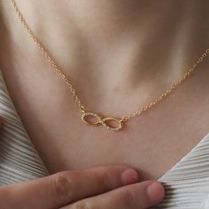 May include: A gold-toned necklace featuring a delicate chain and a central infinity symbol pendant. The infinity charm is embellished with small, clear, sparkling stones set along its curved surface, creating a refined and polished appearance.
