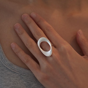 Puede incluir: Un anillo de tono plateado que presenta un diseño grande, liso y ovalado con el centro abierto. El metal tiene un acabado mate y cepillado, creando una superficie suave y no reflectante. La banda del anillo es fina y se une al marco ancho y redondeado.