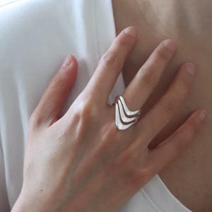 May include: A polished silver ring featuring a multi-layered, wave-like design with three distinct, smooth, curved bands that converge into a single shank. The metallic surface reflects light, highlighting the fluid, sculptural shape of the jewelry.