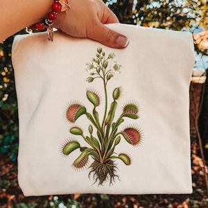 Op de afbeelding: Een crèmekleurig stoffen item met een gedetailleerde botanische illustratie van een Venusvliegenvanger. De plant heeft groene bladeren met roze getinte, stekelige vallen en een centrale stengel met kleine witte bloemen en zichtbare wortels.