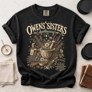 May include: A black short-sleeve t-shirt featuring a cream-colored graphic print. The design displays the text "OWENS SISTERS Apothecary est. 1995" above an illustration of a mortar and pestle, herbs, a glass bottle, and cinnamon sticks.