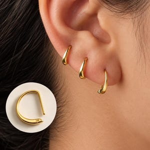 Pode incluir: Três brincos de argola dourados com um design liso e cónico e uma pequena gravação em forma de estrela na extremidade. Os brincos são usados em fila ao longo do lóbulo da orelha e um brinco individual é mostrado separadamente num círculo branco.