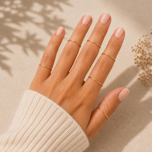 Könnte beinhalten: Ein Set aus sechs dünnen, polierten goldfarbenen Metallringen, die an den Fingern getragen werden. Jeder Ring hat ein schlichtes, glattes und minimalistisches Banddesign. Die Ringe sind über die Finger verteilt, einer am Daumen und fünf an den Fingern.