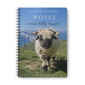 Könnte beinhalten: Ein spiralgebundenes Notizbuch mit einem Walliser Schwarznasenschaf auf dem Einband. Der Text "LITTLE ALPINE NOTES" und "Anna's fluffy thoughts" steht über dem Schaf, während "VALAIS BLACKNOSE - SWISS ALPS" unten vor einer Bergkulisse gedruckt ist.